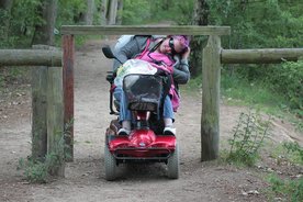 Eine Frau in einem elektrischen Rollstuhl fährt über einen Weg, an dem sie nur sehr schwer weiterkommt durch eine Verengung. 